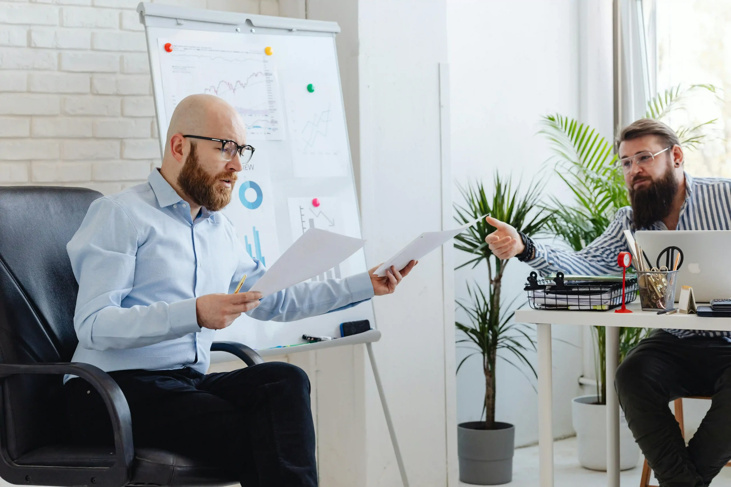 Two businessmen in a discussion, reviewing documents with charts on a whiteboard in a modern office setting.