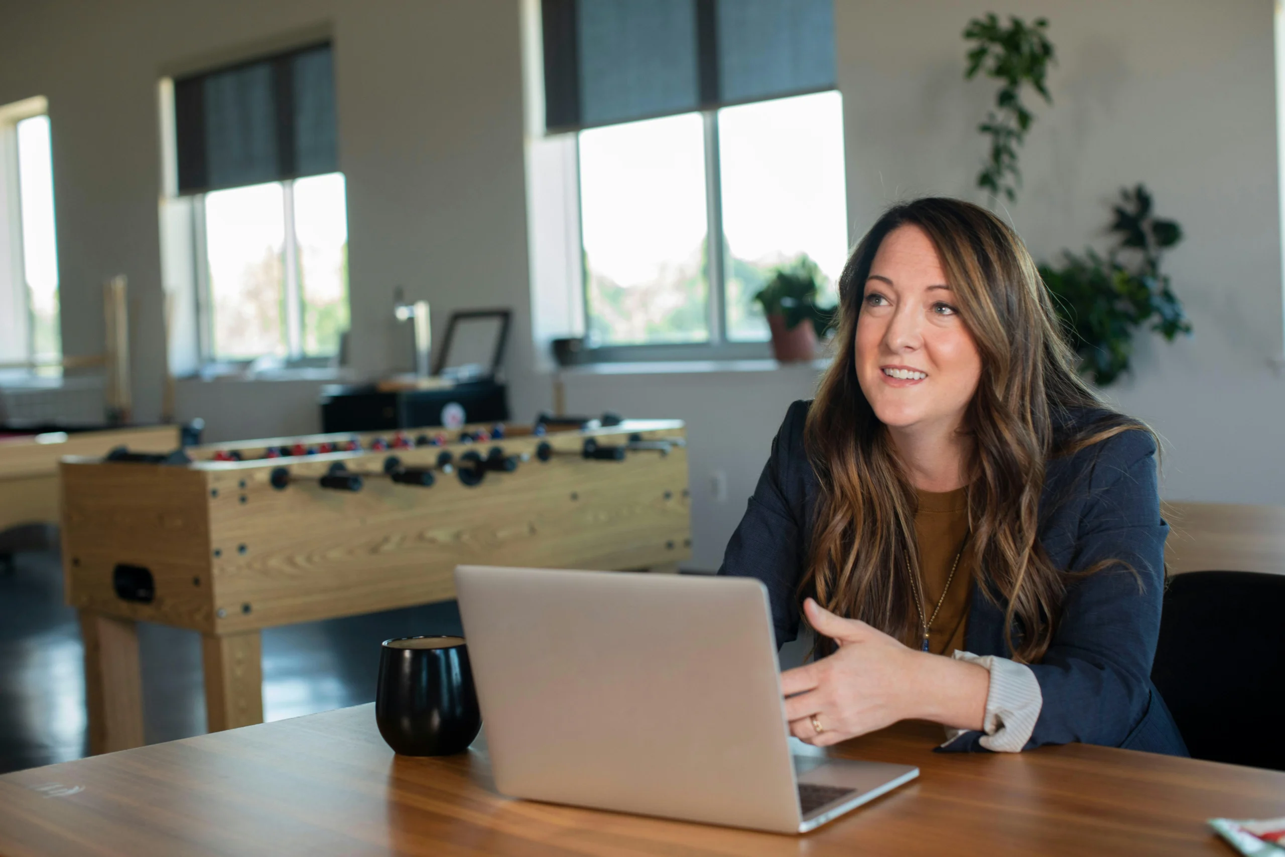 Smiling woman in a blazer sitting at a desk with a laptop in a modern office space.