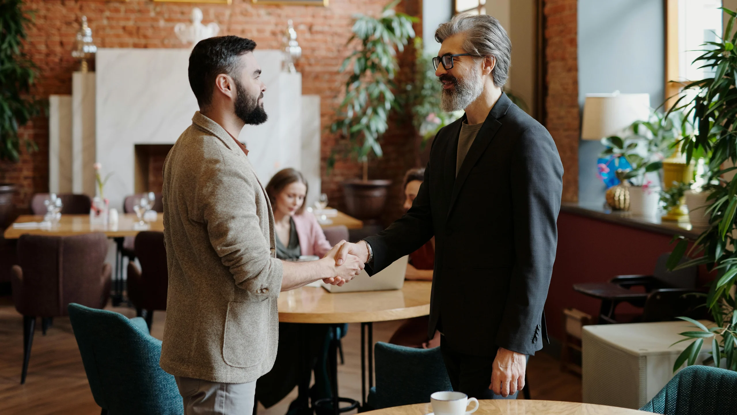 Two professionally dressed men shaking hands in a stylish café with people working in the background.