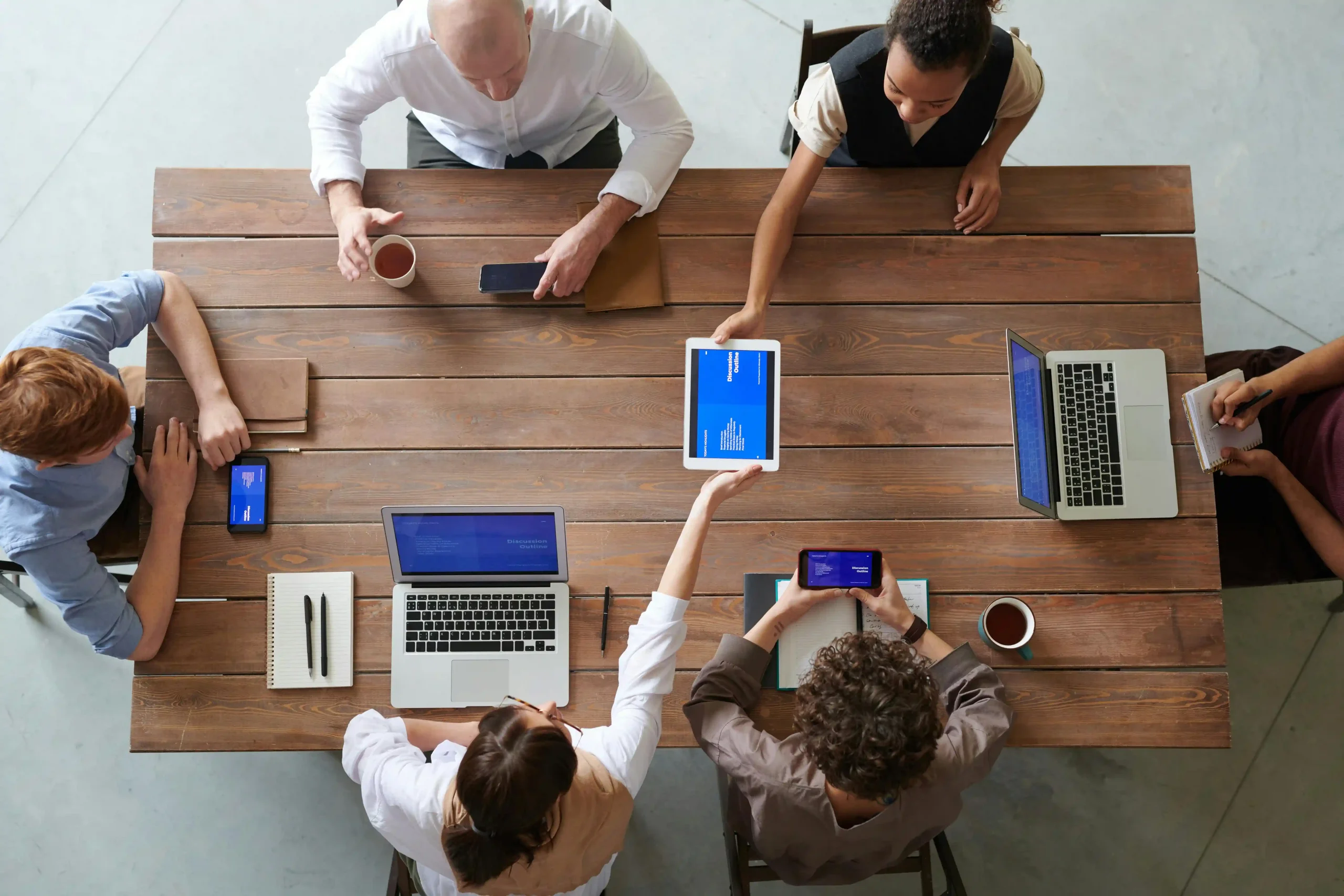 A top-down view of a group of professionals in a meeting, using laptops, tablets, and notebooks around a wooden table.