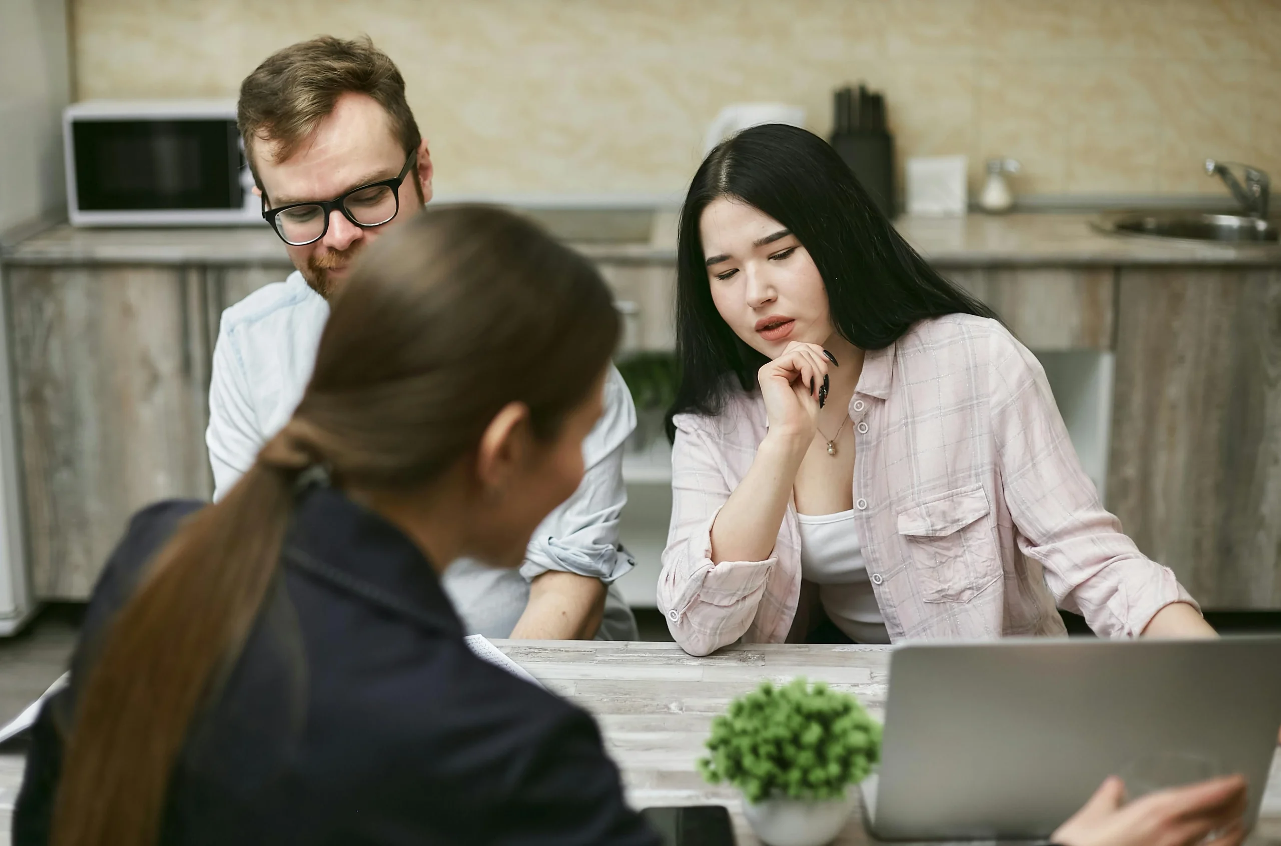 Three people having a discussion at a table with a laptop in a kitchen setting