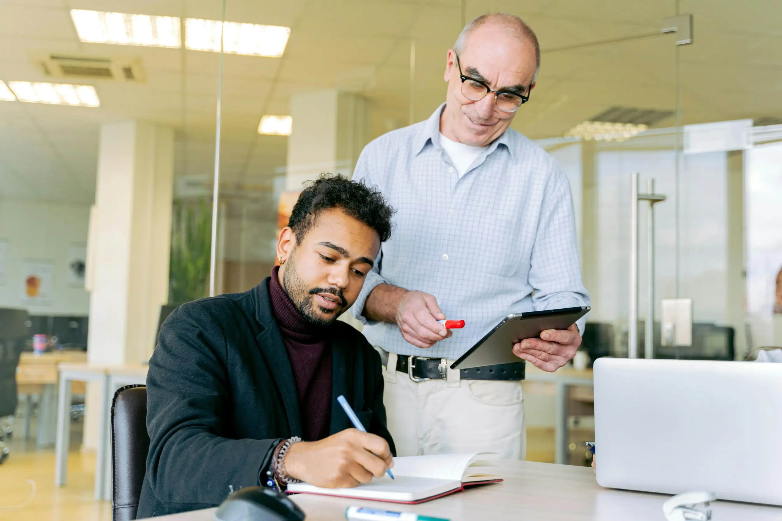 A young professional taking notes while an older colleague reviews information on a tablet in a modern office setting.