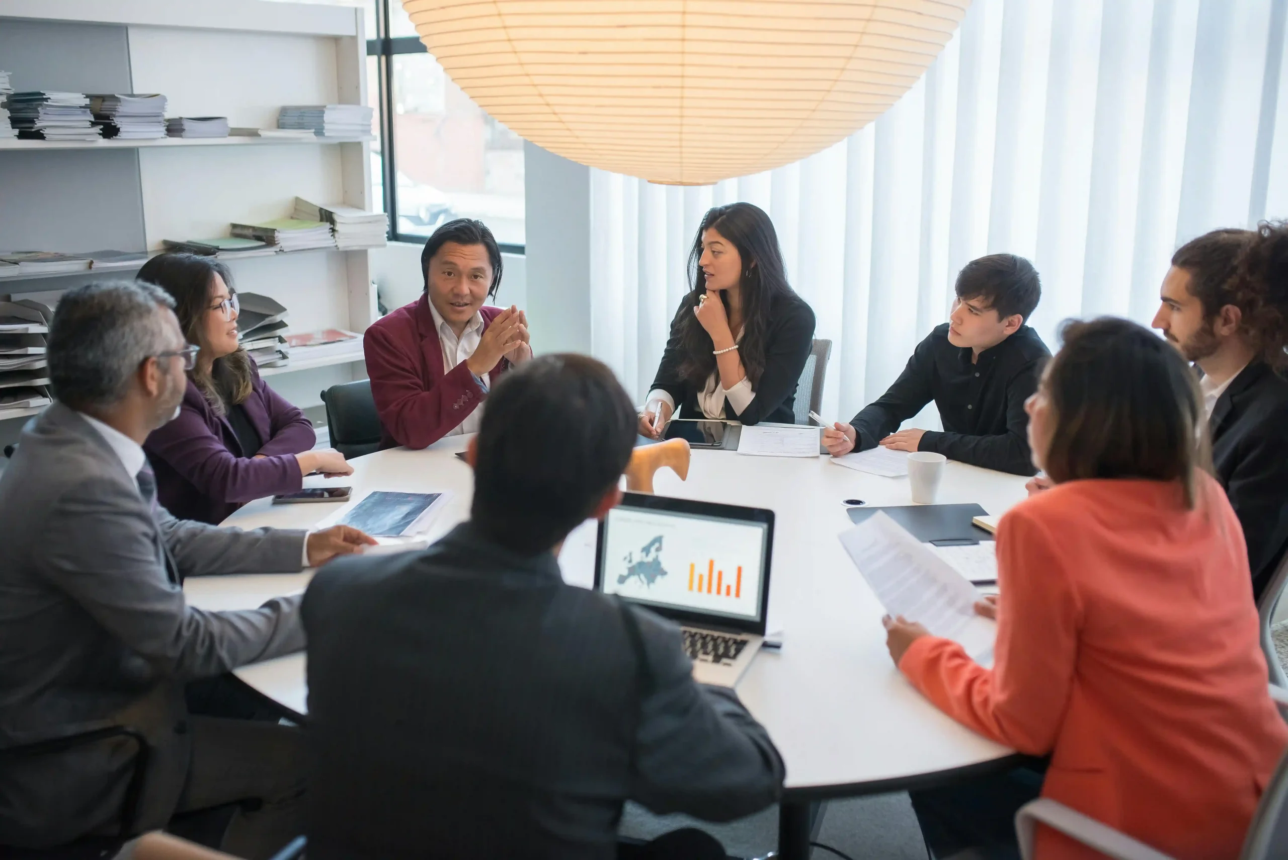 A diverse group of professionals engaged in a business meeting around a conference table, discussing charts and documents.