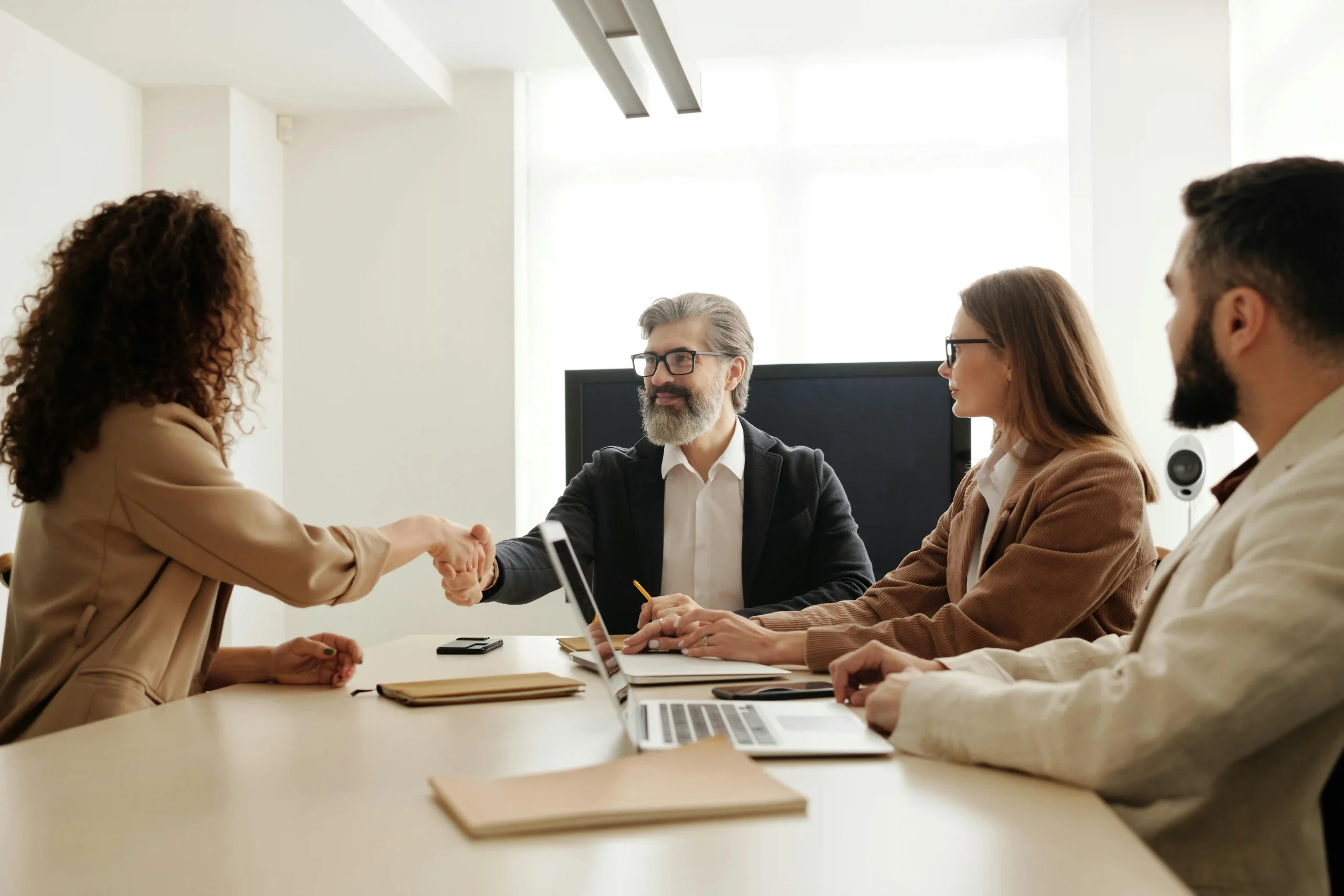 A business meeting where a woman and a man shake hands, with three colleagues observing at a conference table.