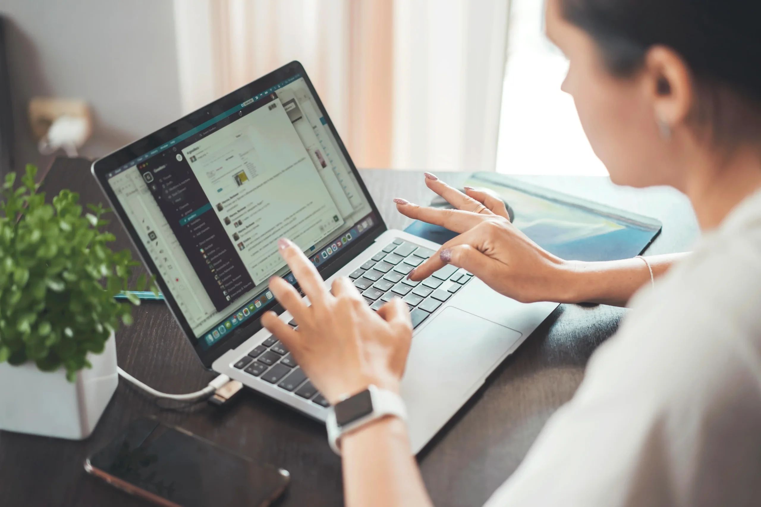 A woman working on a laptop, using a communication platform, with a smartwatch on her wrist and a smartphone nearby.