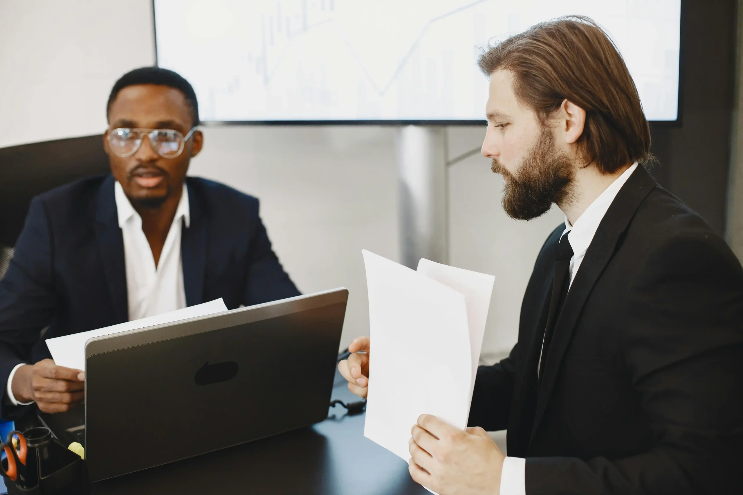 Two businessmen in suits discussing documents in a modern office setting.